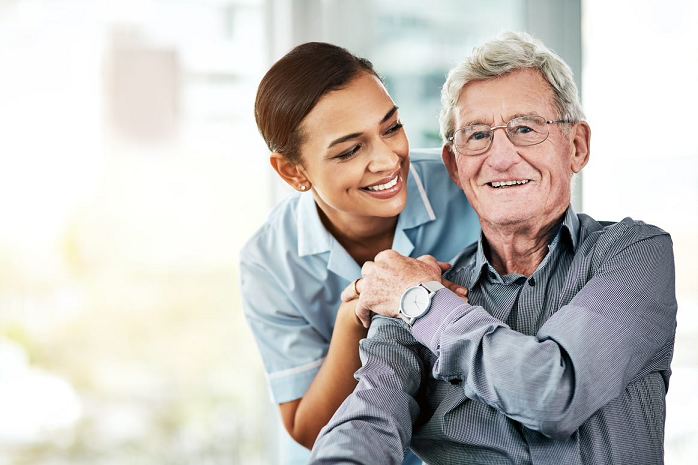 A caretaker leans over an elderly man. She smiles looking at her patient, while he smiles at the camera.