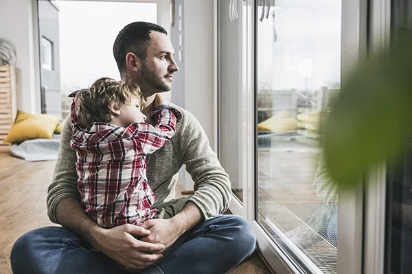 A young father sits cross-legged on the floor and holds his son, who has his arms draped around the dad's neck.